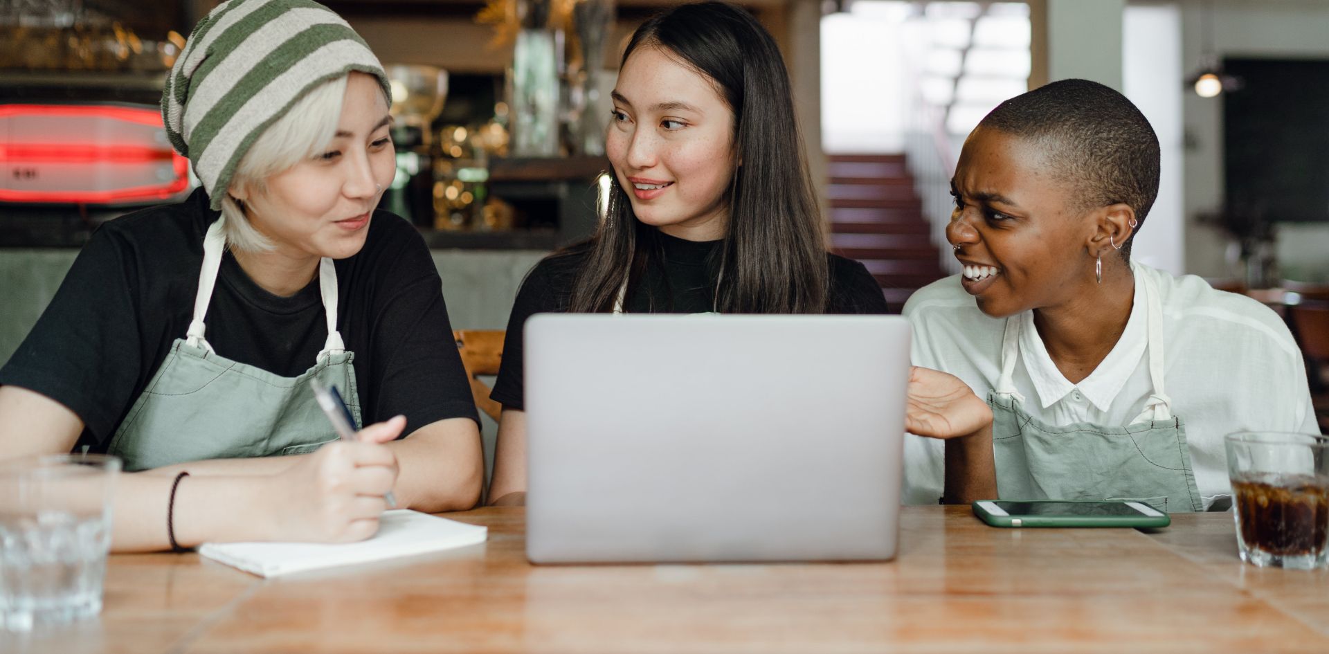 Smiling positive multiracial coworkers wearing aprons browsing modern netbook and having conversation while sitting at wooden table in cozy cafe