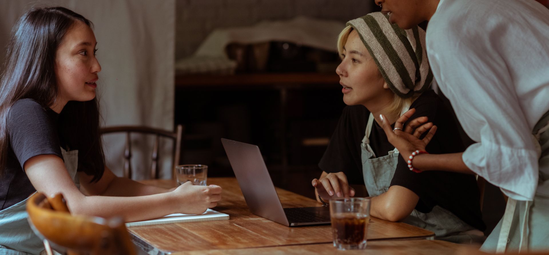 Serious multiethnic waitresses in aprons sitting at wooden table with modern netbook and notebook while discussing business project